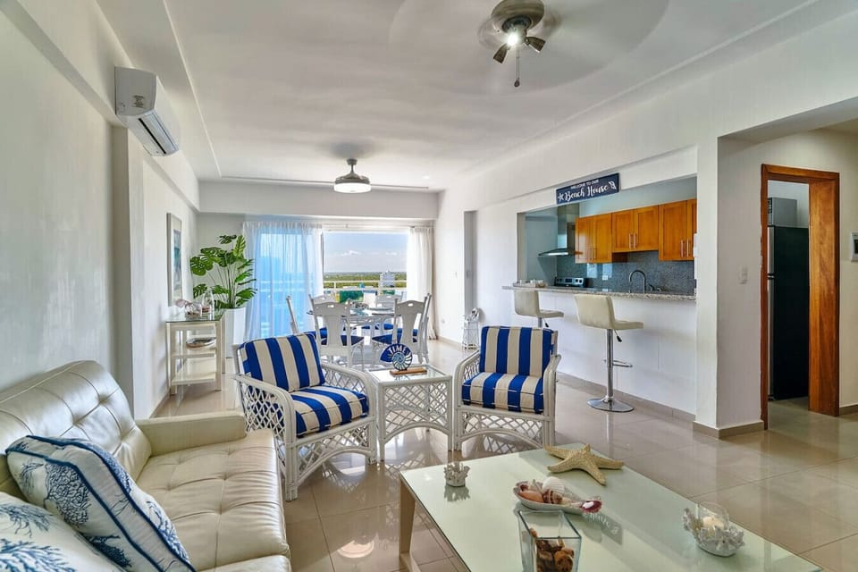 Sala amplia que contiene AC+sofás y en el centro una mesa con decoraciones, al frente se aprecia el área de cocina y desayunador