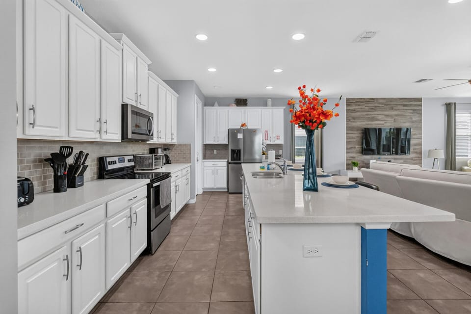 Galley-style kitchen with white cabinetry, beige tile backsplash, stainless appliances, and a quartz island with sink.