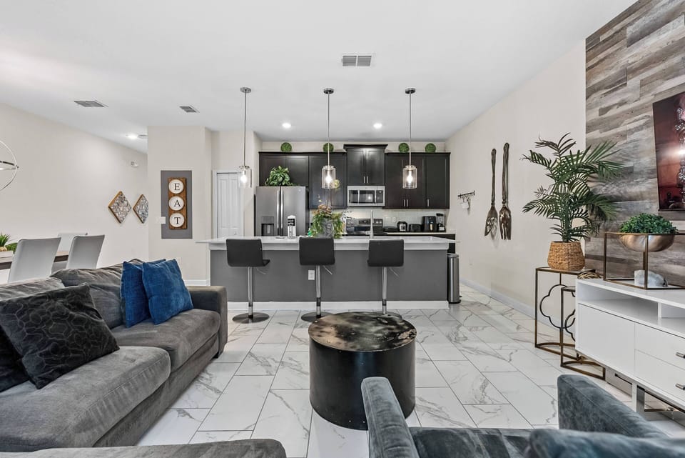 The open layout connects the chic kitchen with stylish bar seating to a cozy living area with plush pillows and a sleek marble floor, creating a warm and welcoming ambiance with natural textures and rich cabinetry tones.