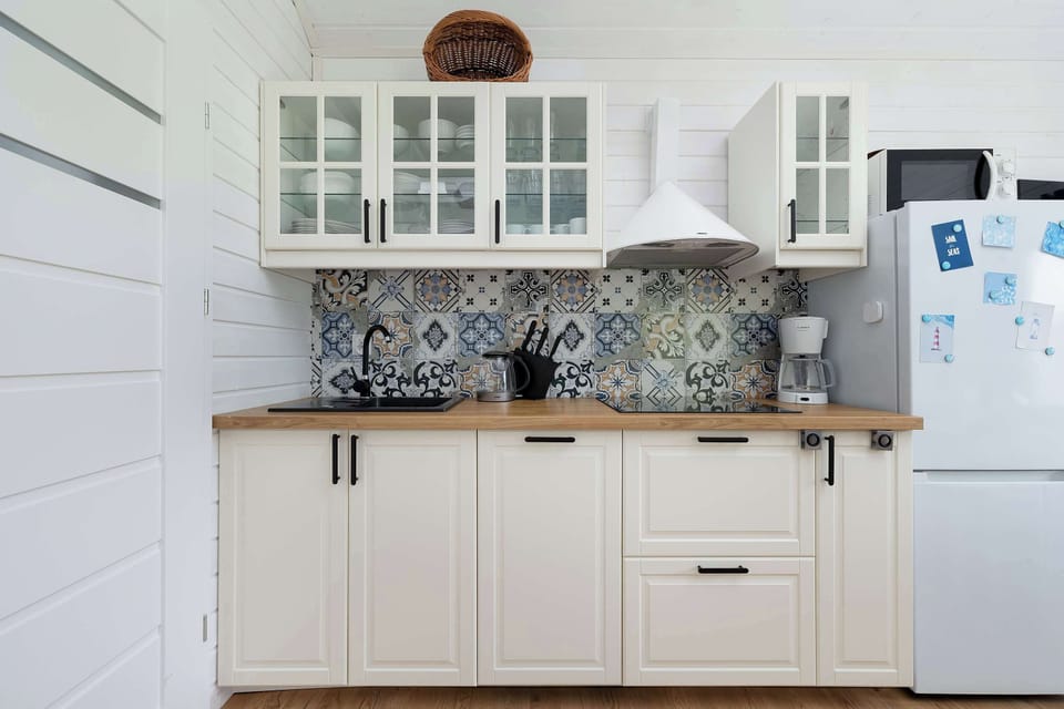 A compact kitchenette with white cabinets and patterned backsplash tiles. A fridge and open shelves complete the space.
