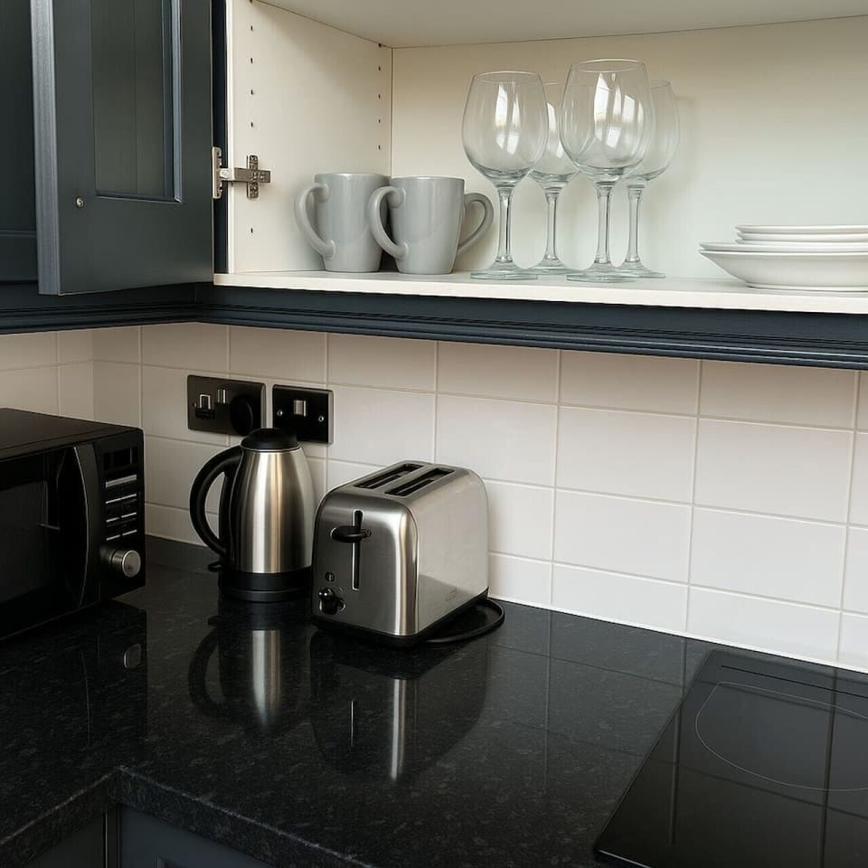 Modern kitchen corner with kettle, toaster, microwave, and electric hob. Cupboard stocked with mugs, wine glasses, and plates. Clean black countertop and white tiled splashback for a fresh, welcoming feel.