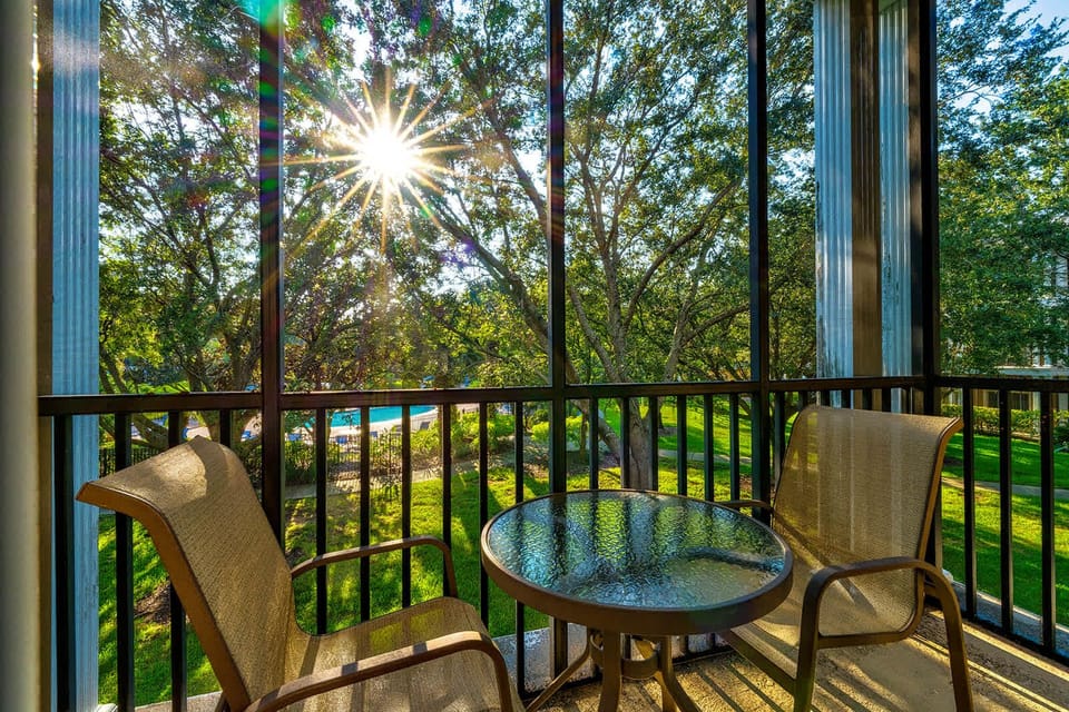 Screened balcony with seating overlooking a sunlit garden and pool area.