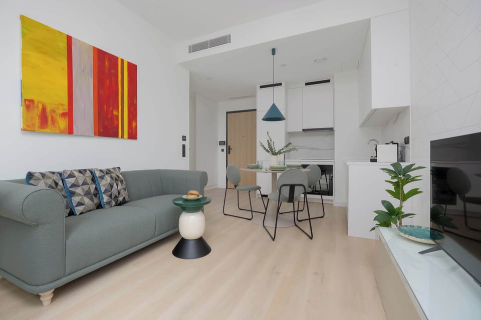 A bright living room with a sofa, dining table, and large windows letting in natural light. The space feels modern and inviting, ideal for relaxation or shared meals.
