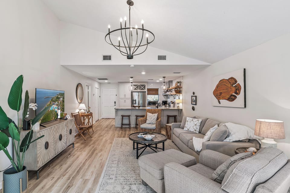 Sink into plush gray sectional seating in your open-concept living space, where modern coastal elegance meets comfort beneath soaring vaulted ceilings and warm chandelier light.