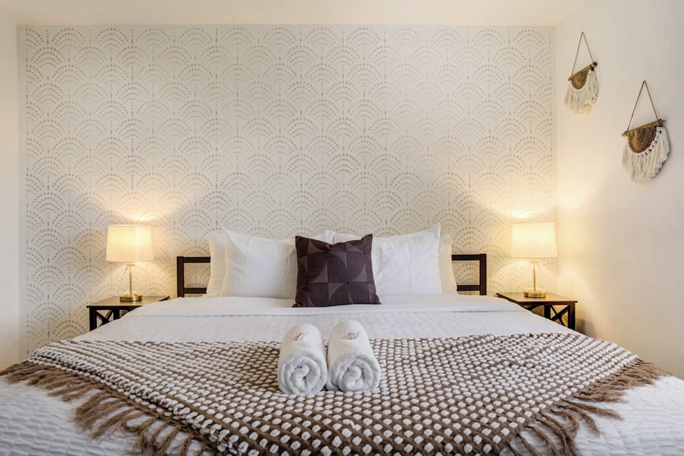 Spacious king bedroom featuring a soft patterned accent wall, warm bedside lighting, and large windows that let in natural light with views of the surrounding trees. A comfortable retreat designed for relaxation.