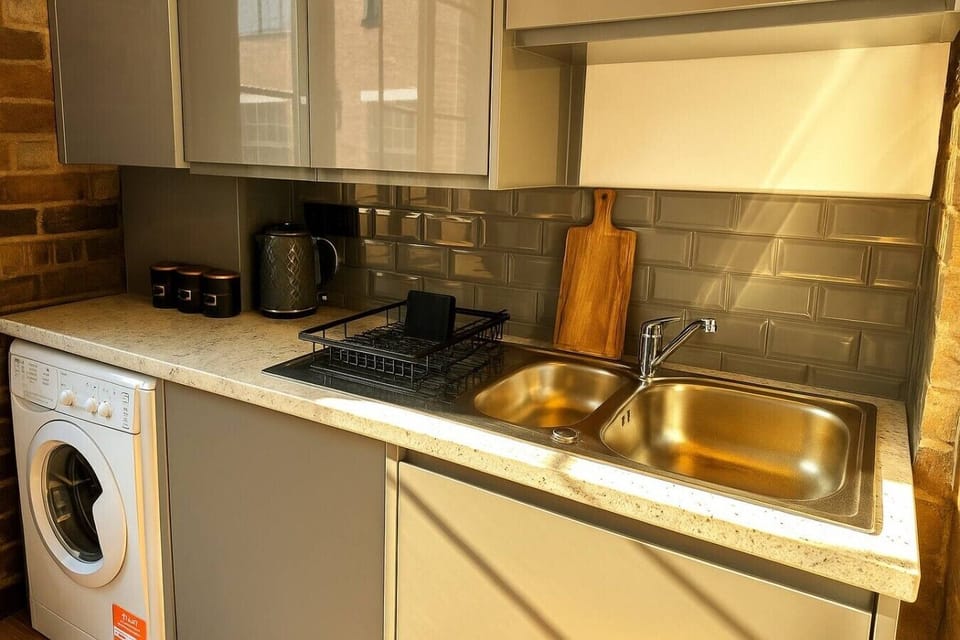 Compact and practical kitchenette featuring a stainless steel sink, modern backsplash, washing machine, kettle, and storage cabinets. A functional space designed to make your stay comfortable and convenient with all essentials close at hand.