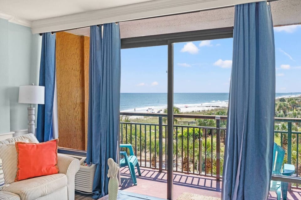 A bright living room with coastal décor and a comfy sofa sleeper, opening to a private balcony. Large windows frame stunning ocean and pool views, filling the space with natural light and a relaxing beachside atmosphere