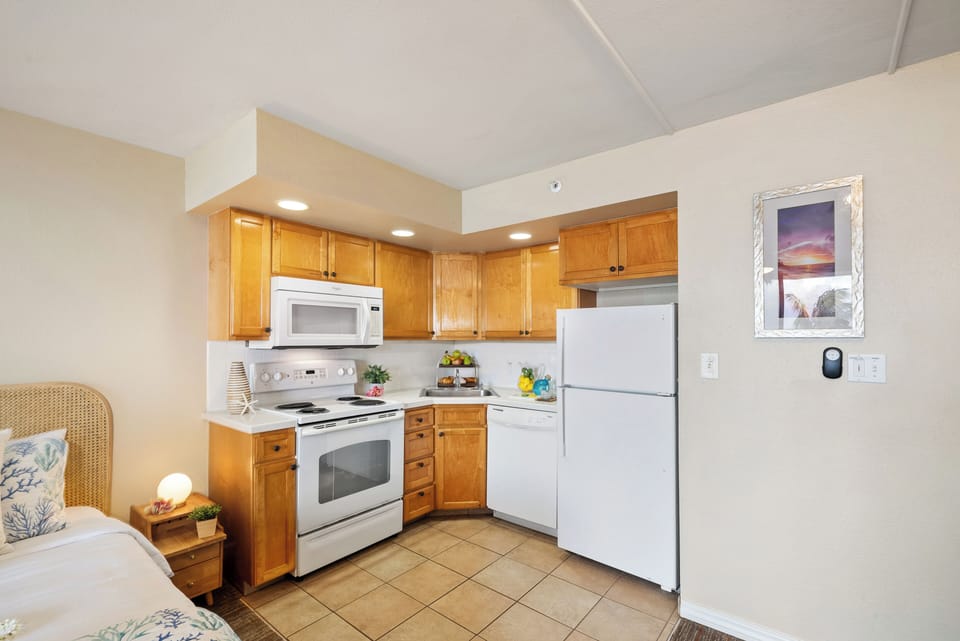 Kitchen with light brown wooden cabinets, white microwave, stove and refrigerator, beige tiled flooring and countertop, decorative fruit bowl adding warmth.