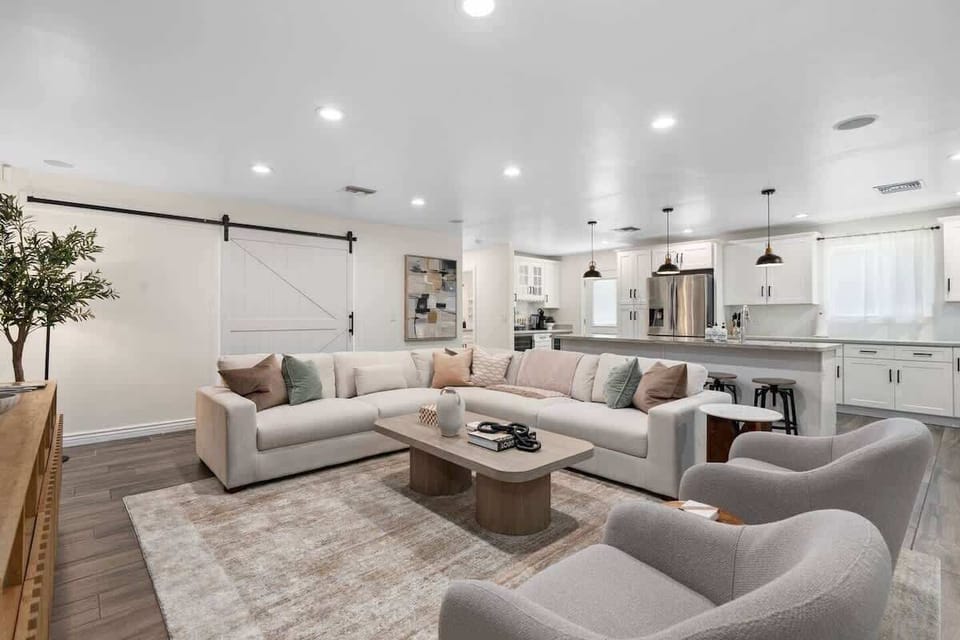 Bright open living area with a roomy sectional, soft rug, and cushy chairs made for movie nights. Set snacks on the wood coffee table, then drift to the kitchen island where stools line up under pendant lights. Barn door detail by the sofa.