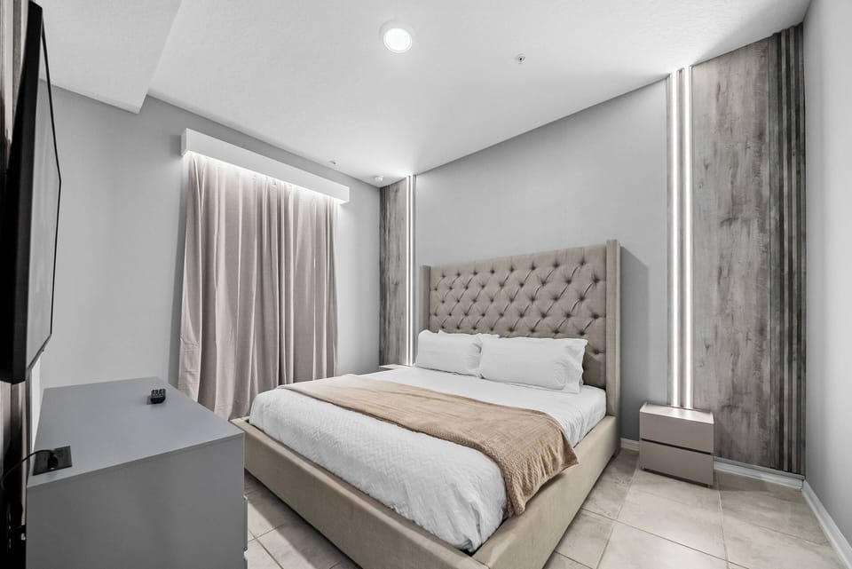 Experience a stylish bedroom with a plush tufted headboard and a soothing neutral palette, complemented by soft lighting and sleek textures. The large window drapes invite natural light, making your stay warm and inviting.