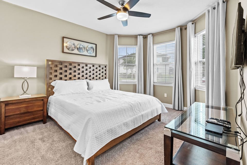 Experience this thoughtfully designed bedroom with a cozy king bed, stylish headboard, and soft beige carpeting. Tall windows adorned with light curtains invite natural light, creating a warm and inviting atmosphere for your stay.