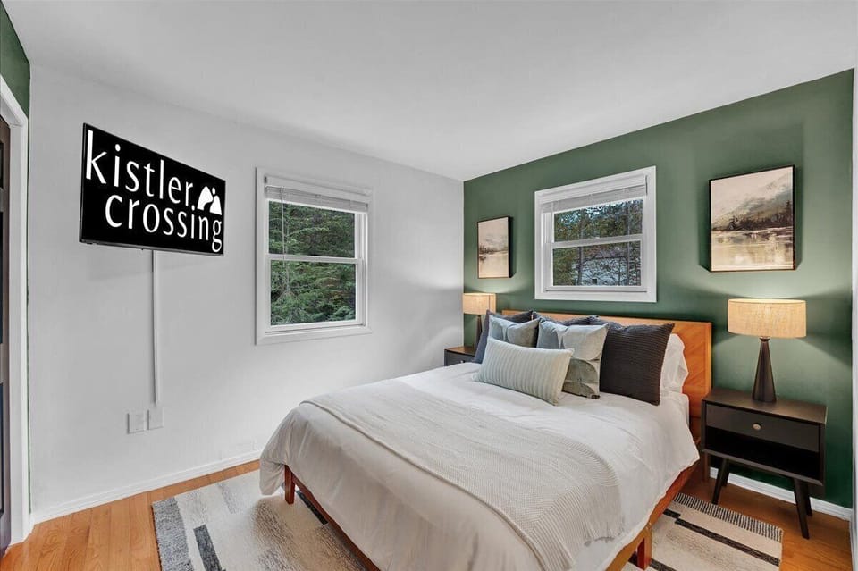 Unwind in this serene bedroom sanctuary. A deep green accent wall complements the cozy bed with crisp white linens. Warm light from modern lamps and large windows with tranquil forest views create a perfect, peaceful escape.