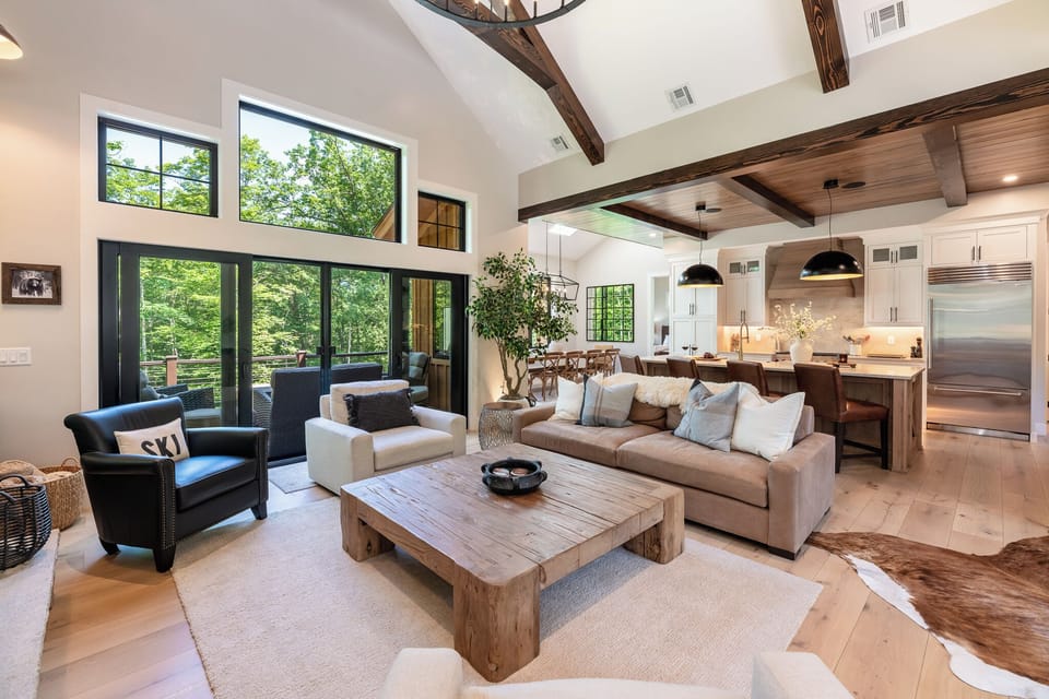 Inviting living room with vaulted ceilings, exposed beams, and large windows framing scenic views. Cozy seating flows seamlessly into the kitchen and dining area, creating the perfect open-concept gathering space.