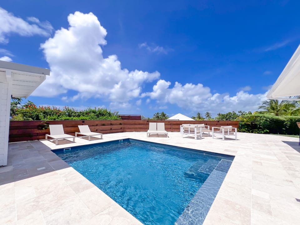 Modern pool surrounded by sunbeds and outdoor furniture, under a tropical blue sky with scattered clouds.