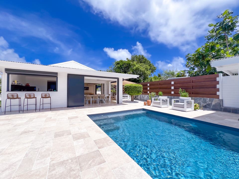 Elegant pool surrounded by stone terrace, with pool house, bar and lounge areas under the tropical blue sky.