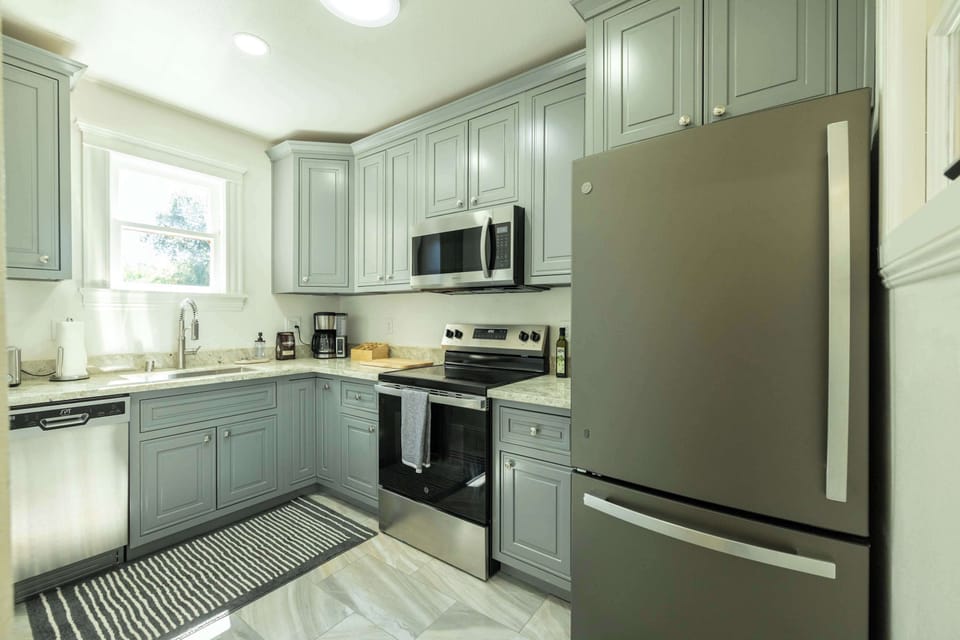 Cook with ease in this bright, fully equipped kitchen featuring modern stainless steel appliances, soft sage cabinetry, and a sunny window that fills the space with natural light