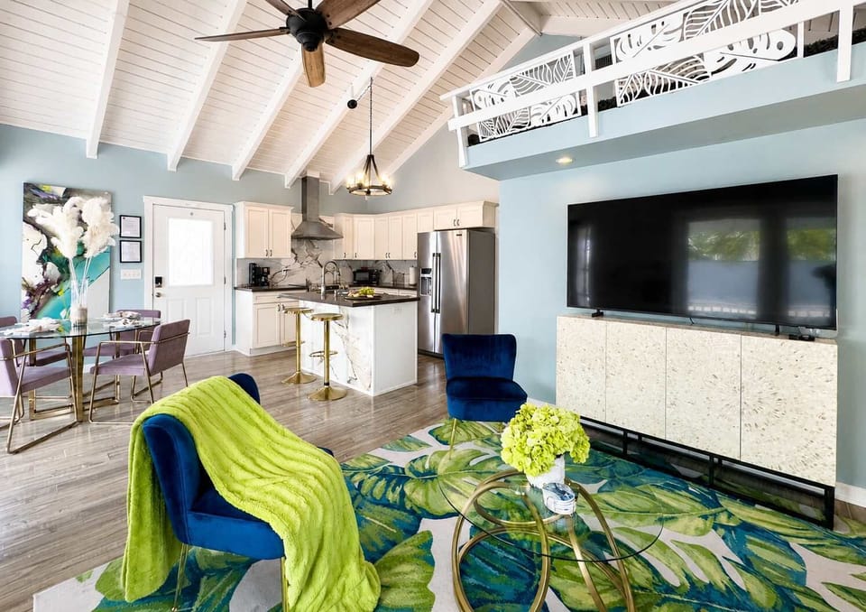 Inviting open-concept living area with soaring ceilings, modern coastal décor, and a large flat-screen TV — seamlessly connected to the fully equipped kitchen and dining space for easy entertaining.