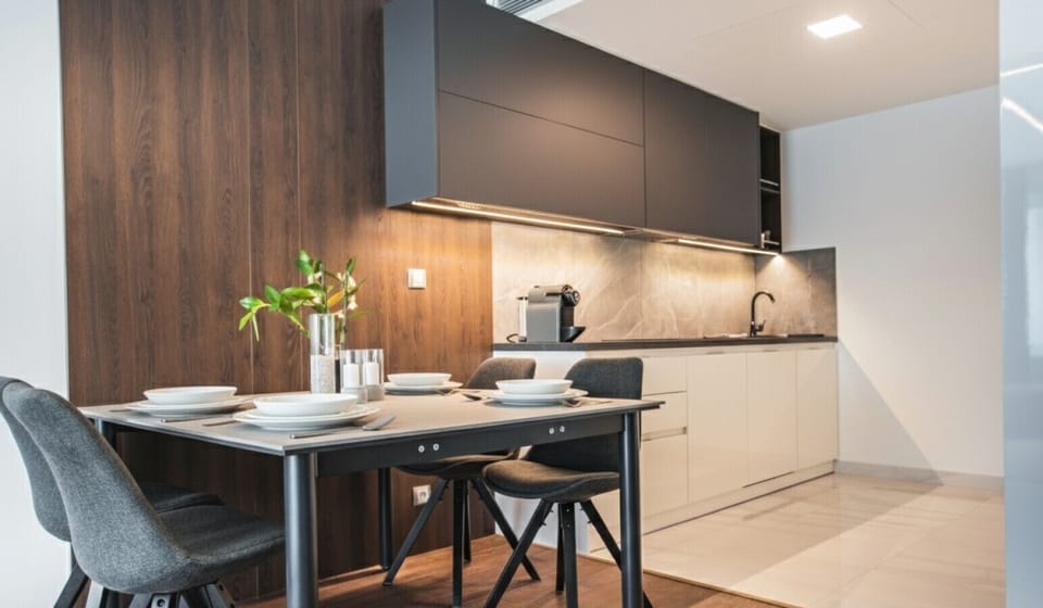 Modern kitchen with dark wood accents, sleek cabinets, and a dining area. The table is set with two chairs and a small plant centerpiece. The kitchen features a built-in oven, a sink, and ample counter space.
