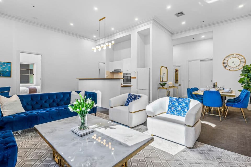 Spacious open-plan living area with plush blue seating, marble coffee table, and elegant dining space. Bright lighting, modern kitchen, and stylish decor create a perfect setting for relaxing or entertaining.