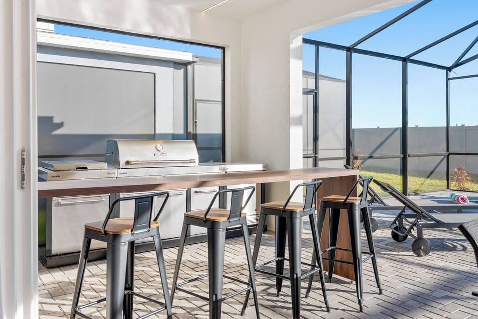 Enjoy alfresco dining in our stylish outdoor kitchen, complete with sleek metal barstools and a spacious wooden countertop. The abundant natural light from large windows sets a vibrant mood, ideal for gatherings.