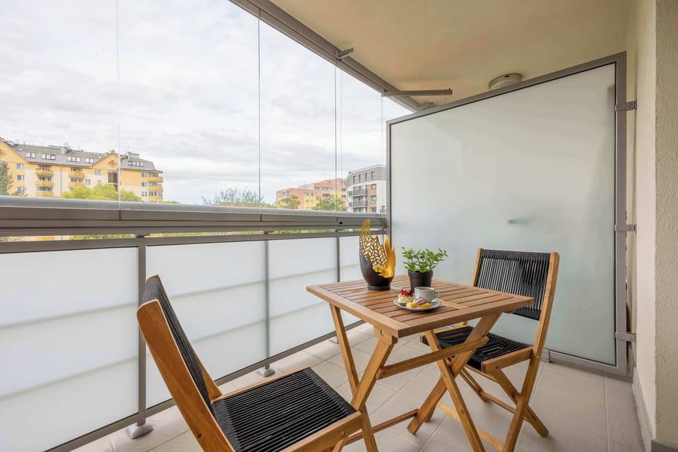 A small balcony with a table and two chairs overlooking nearby buildings. A pleasant spot for enjoying coffee or meals outdoors.