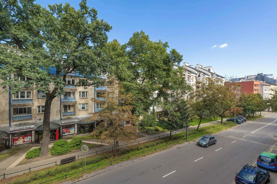 A street view of the apartment building with trees lining the road. Vehicles are parked along the street, giving a sense of the neighborhood.