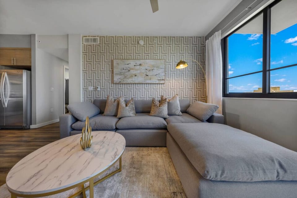 Cozy Lounge with Natural Light-Comfortable living area featuring a plush sectional sofa, chic décor, and a marble-style coffee table. Large windows bring in plenty of sunlight and city views.