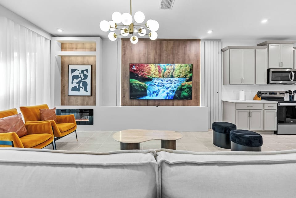 This bright living room showcases beautiful artwork above a cozy light gray sofa and vibrant orange chairs. A modern chandelier adds warmth, while a calming nature view on the TV brings a touch of tranquility to the space.