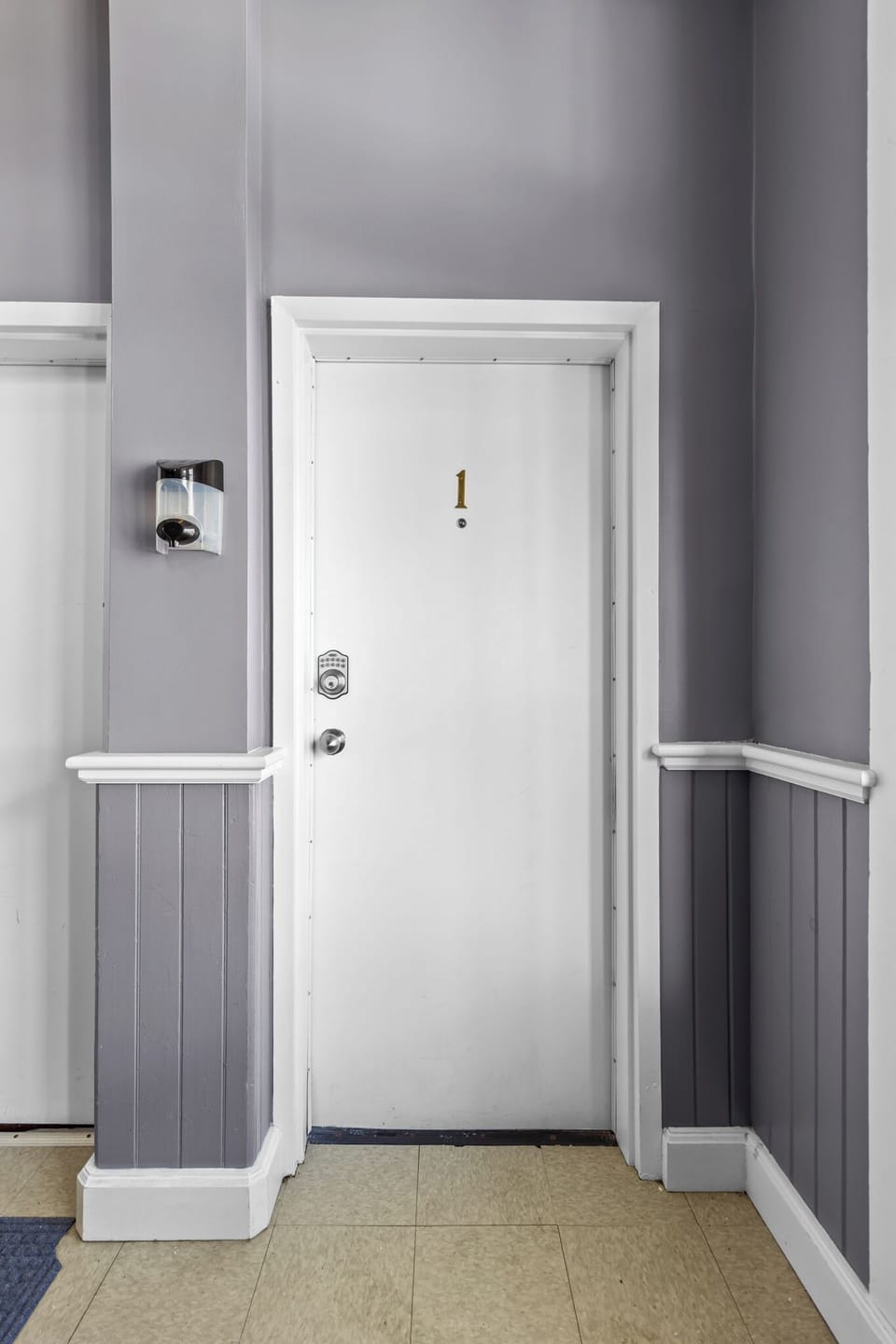 Private and secure apartment entrance marked clearly as Unit 1. The entryway features modern grey wainscoting and a bright white door, providing a clean and welcoming first impression for arriving guests.