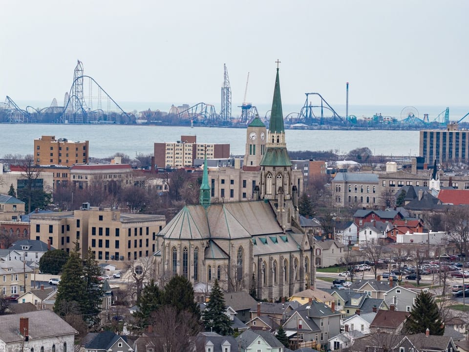 Historic cityscape featuring a prominent church with green spires, surrounded by diverse architecture and waterfront amusement park rides in the distance.