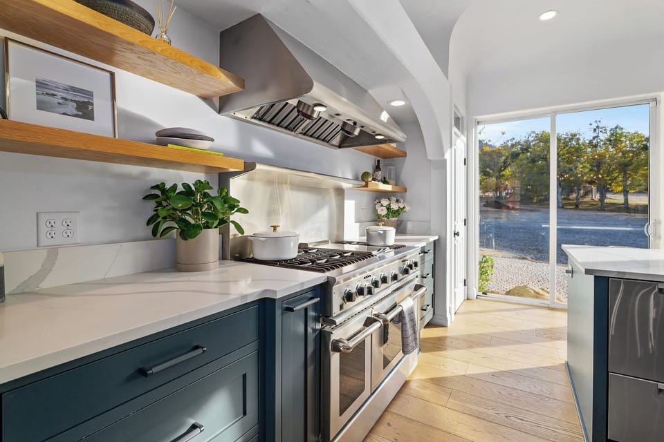 Every detail designed for style and function. We love the rich blue cabinetry contrasted with the crisp white countertops and the warmth of the floating wooden shelves—all centered around this high-end stainless steel range.