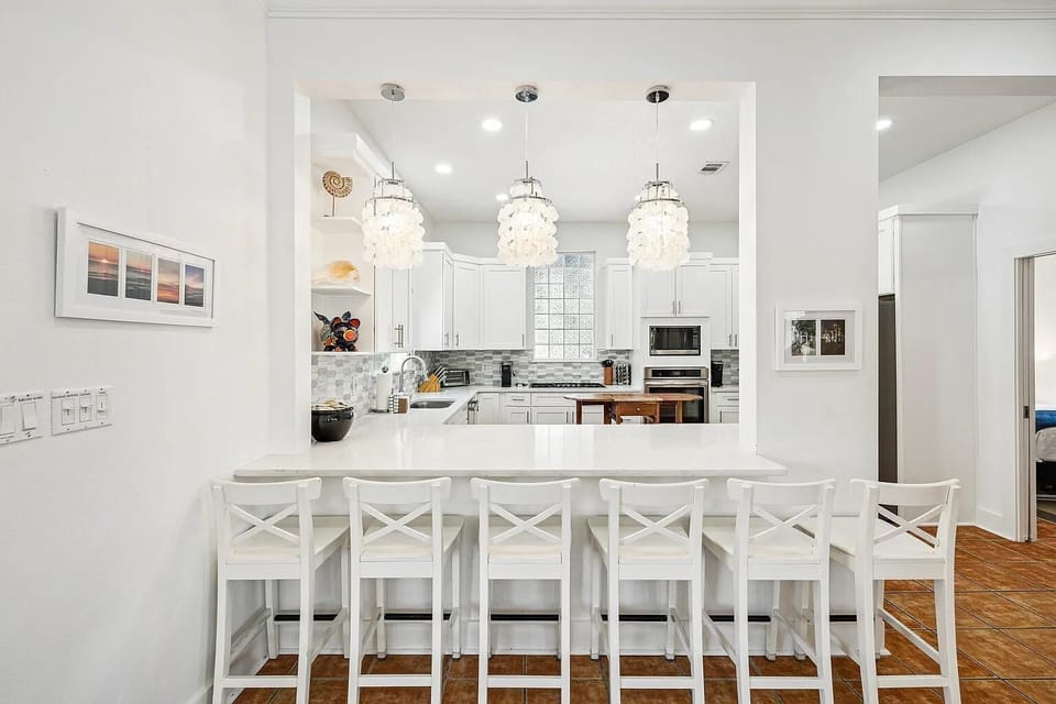 White breakfast bar with six cross-back stools faces a kitchen with marble countertops and pendant lighting.