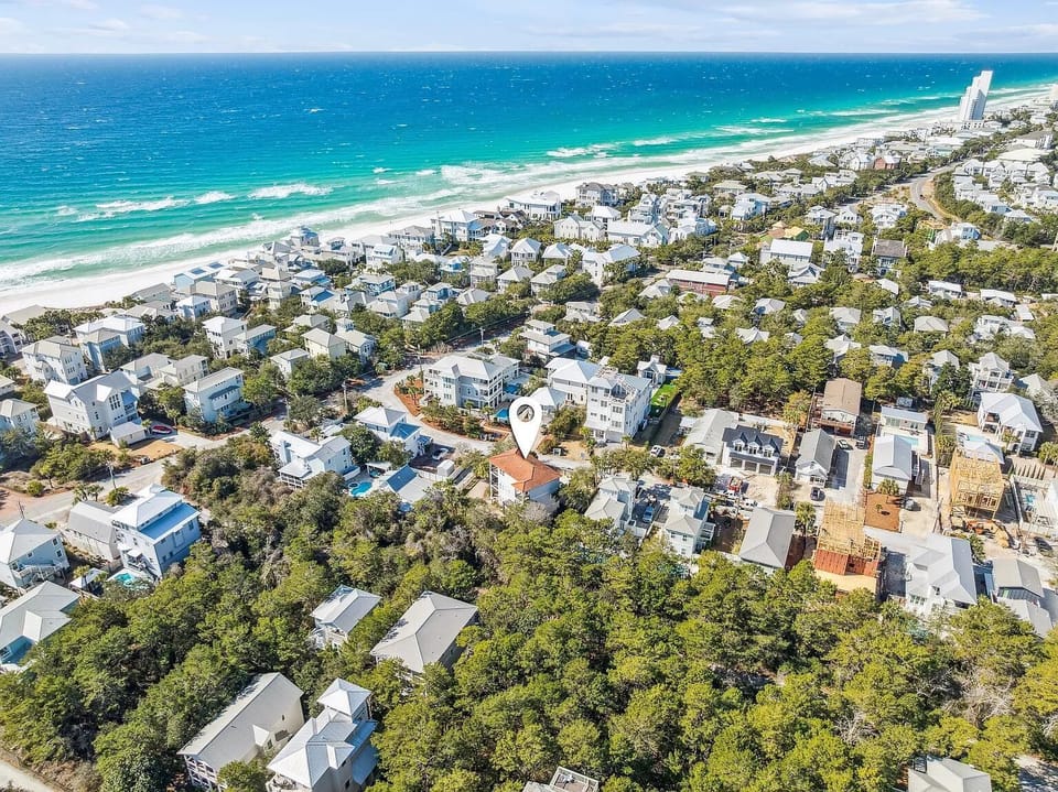 Aerial view of a coastal neighborhood with white and gray homes, green trees, and the turquoise ocean shoreline.