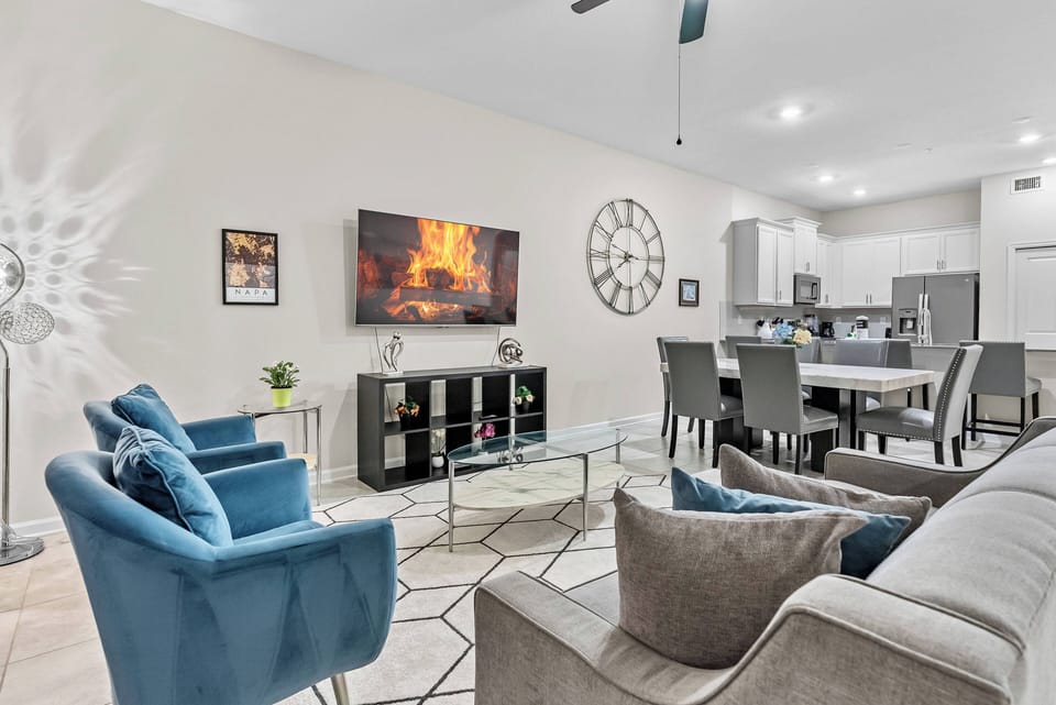 Enjoy a bright, spacious living area with cozy gray sofas and plush blue accent chairs, perfect for relaxing. The stylish coffee table and striking wall clock enhance the open layout that connects seamlessly to a modern kitchen.