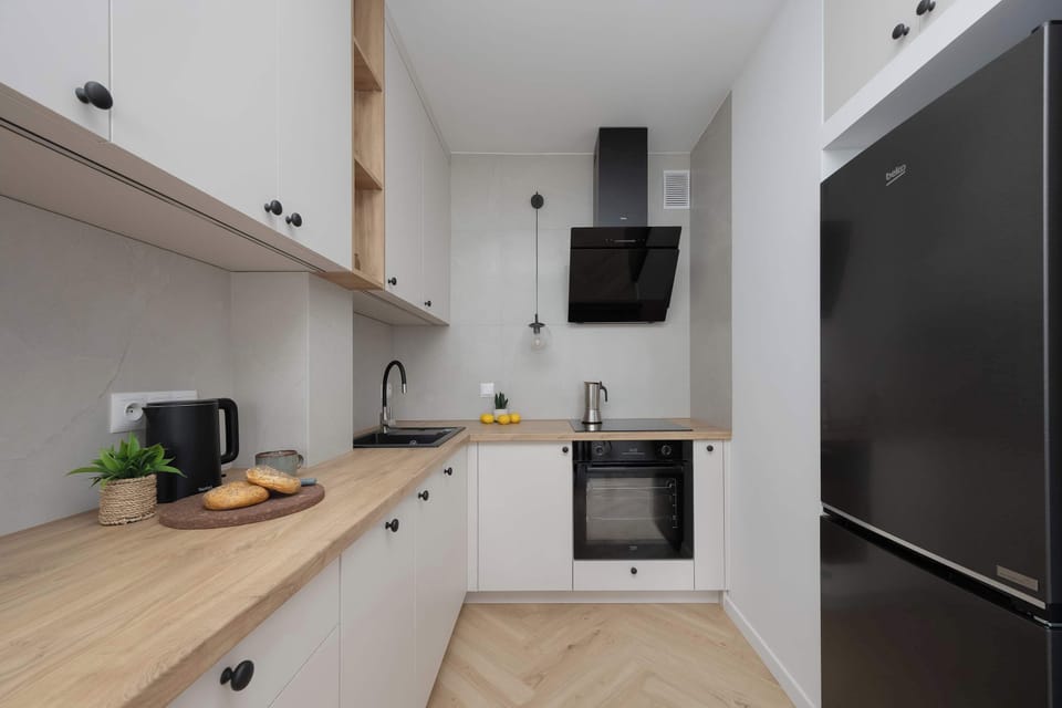 A modern kitchenette with white cabinets, a black oven, induction cooktop, and a tall fridge. The space is compact yet fully functional for cooking.