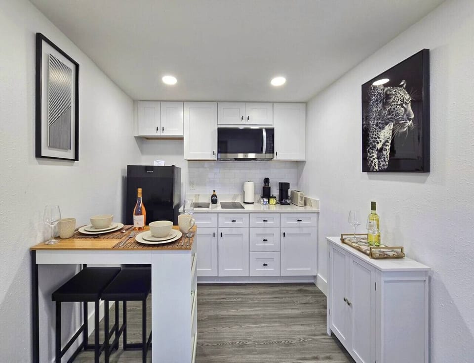 This small, modern kitchenette features white cabinets, a subway tile backsplash, and stainless steel appliances. Recessed lighting illuminates the space. The kitchen includes a breakfast bar with two stools, set for a meal, and artwork of a leopard and a