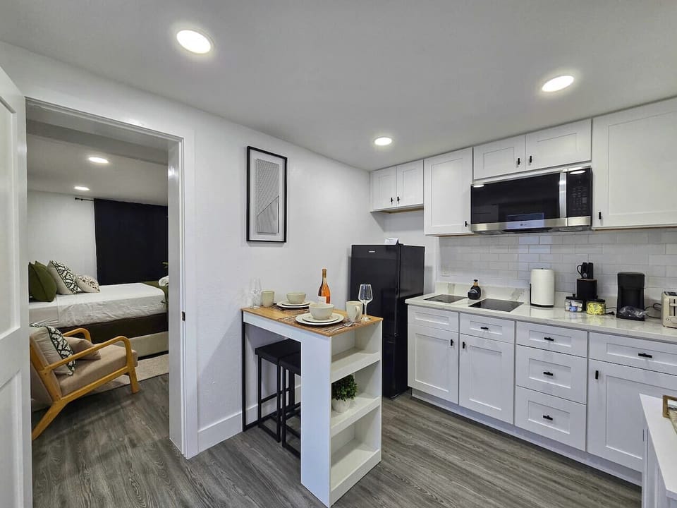 Modern, fully equipped kitchen with sleek white cabinets and counters. Includes a refrigerator, microwave, double induction cooktop, and dining area for two. Direct access to the bedroom.
