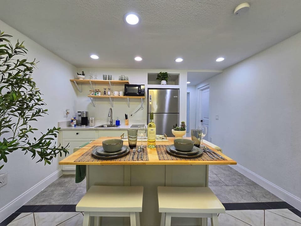 This interior shot features a kitchenette/dining area with a light-wood topped island set for two with gray dishware. The kitchen has white lower cabinets, a stainless steel fridge, and a microwave. Floating shelves hold glasses and decor. Recessed lighti