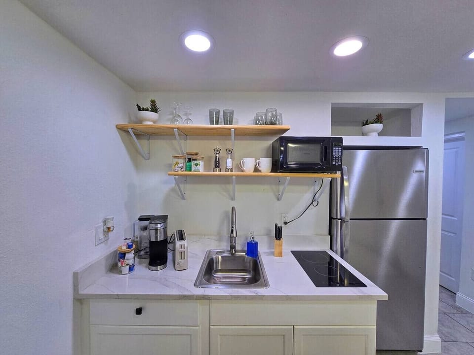 This modern kitchenette features a stainless steel refrigerator, a built-in microwave, and a white quartz-look countertop with an undermount sink and induction cooktop. Two wooden floating shelves display glassware and small decor. Appliances include a co