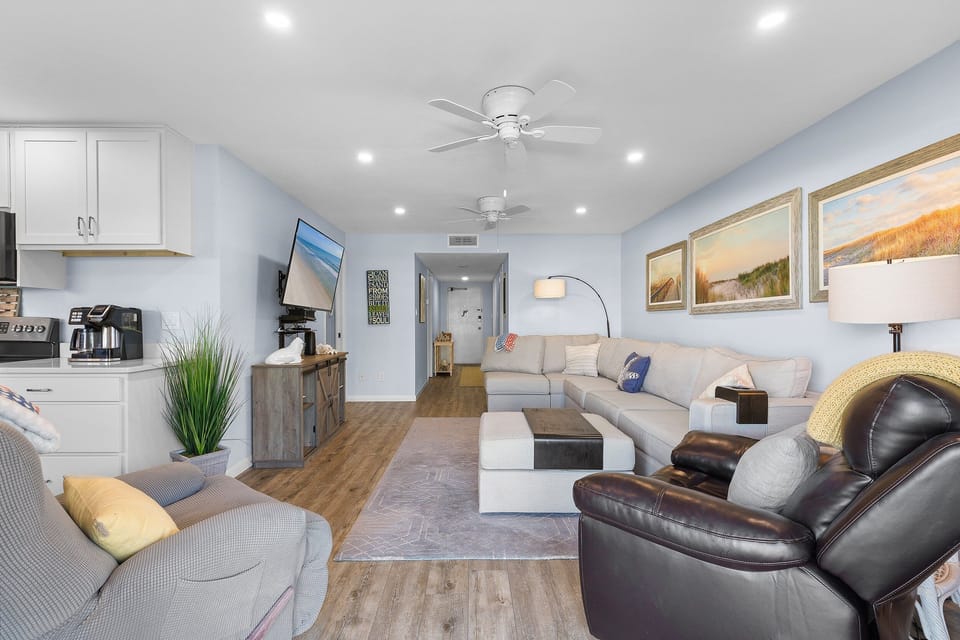 Unwind in this airy coastal living space featuring plush seating, beach décor, and an open layout that flows into the kitchen—perfect for relaxing after a day of seaside adventures.