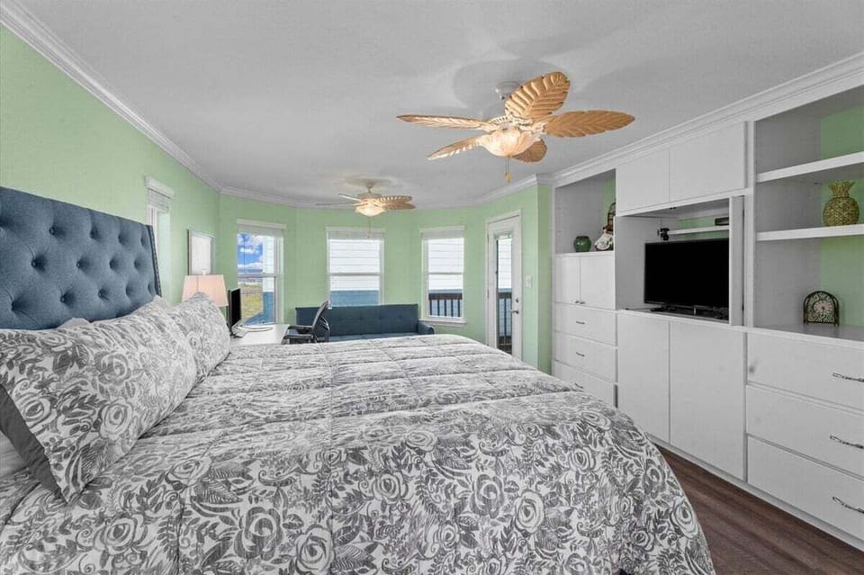 Bedroom with large bed, built-in storage, TV, ceiling fan, and soft coastal green walls for a relaxing stay.