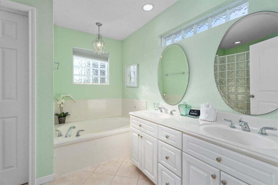 Spacious bathroom with double vanity, soaking tub, large mirror, soft green walls, and bright natural light.