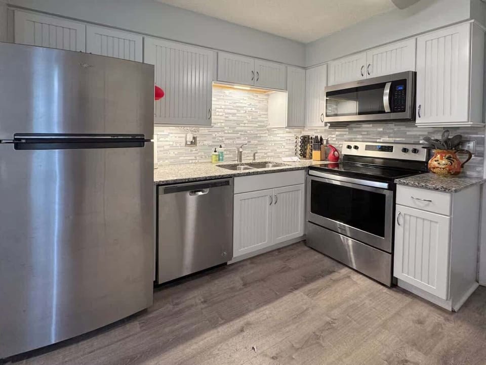 A fully equipped kitchen with clean white cabinetry, granite countertops, and a chic tiled backsplash. High-end stainless steel appliances include a full-sized refrigerator, dishwasher, oven/range, and microwave.