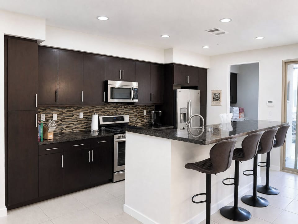 Your fully-equipped modern kitchen features sleek dark cabinetry, granite countertops, stainless appliances, and a breakfast bar with four stylish stools—perfect for preparing meals or enjoying morning coffee together.