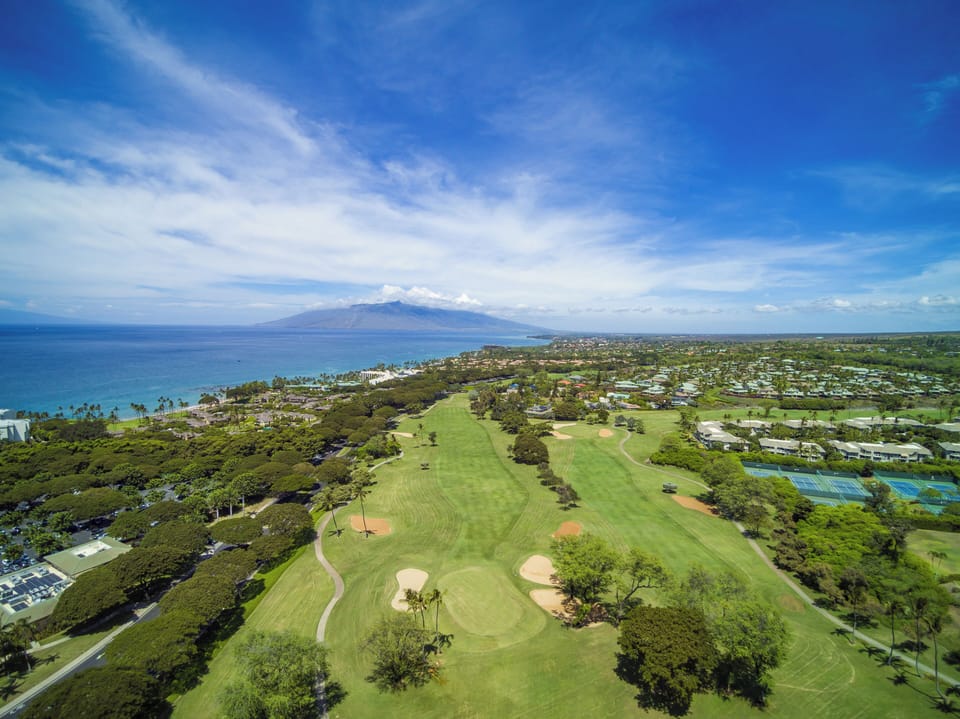 Aerial view of the surrounding coastal area featuring a championship golf course, residential neighborhoods, and ocean views with mountain backdrop.