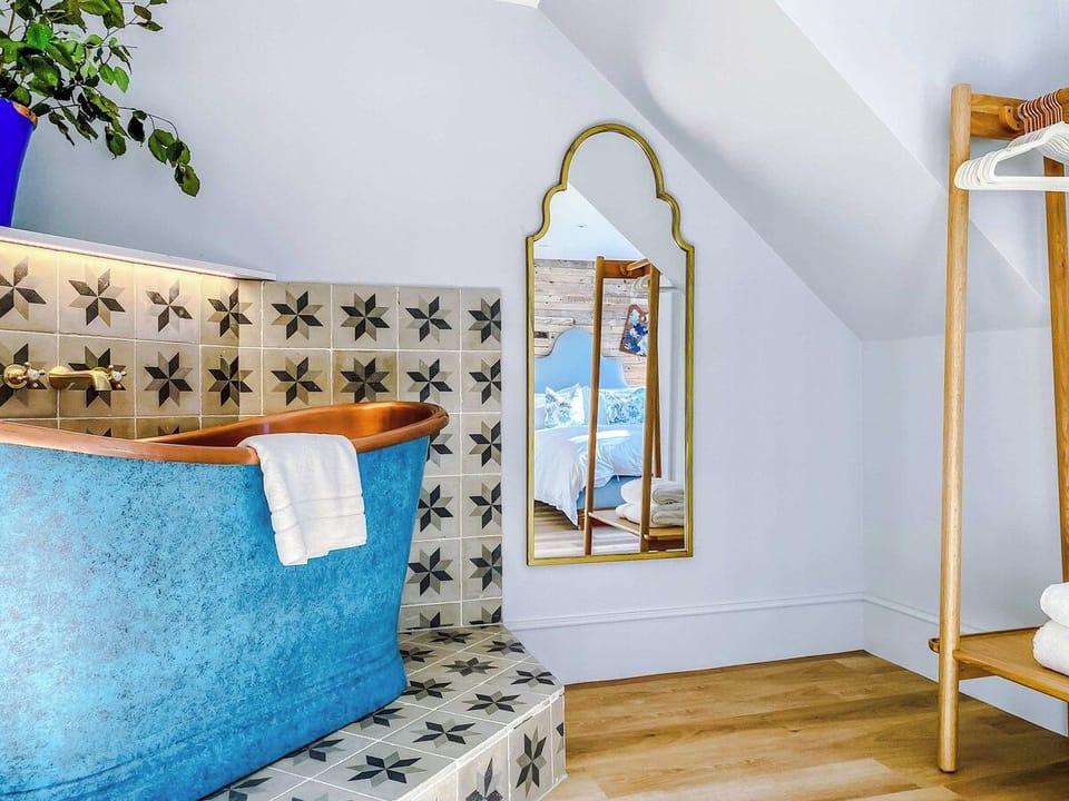 Stylish bathroom with a freestanding blue tub and copper rim set against patterned tiles. A recessed shelf with greenery, soft lighting, and a gold-framed mirror add elegance, while natural light creates a cozy, inviting retreat.