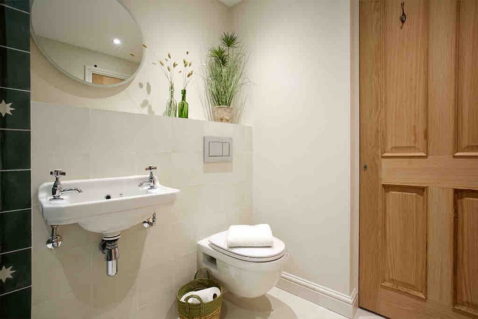 Stylish bathroom with a wall‑mounted sink, round mirror, and modern toilet. Light walls, wicker accents, and greenery bring warmth, while clean lines and soft lighting create a contemporary, welcoming retreat.