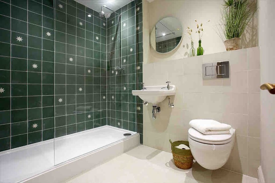Modern bathroom with a walk‑in shower featuring floral green tiles, sleek wall‑mounted sink, and round mirror. Warm lighting, neutral tones, and stylish décor create a clean, contemporary retreat with comfort and charm.
