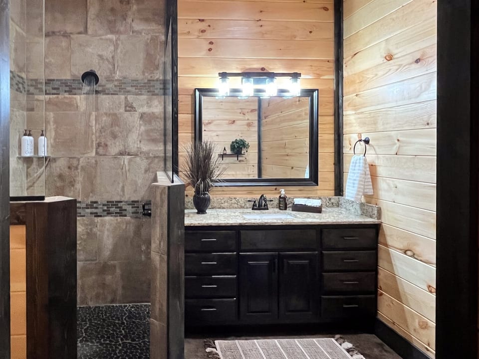  A well-designed bathroom featuring a granite vanity, rustic wood walls, and a large mirror. The open walk-in shower adds a touch of luxury.