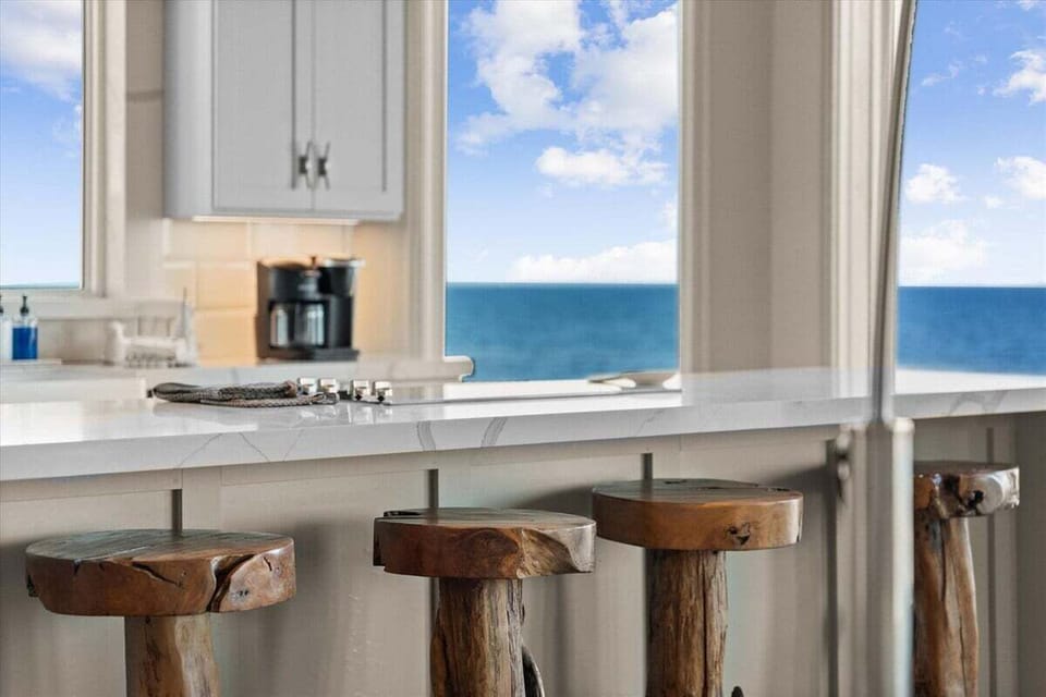 Hand-carved wood stools line the marble island, their natural grain echoing the textures of driftwood and sand beyond the window.